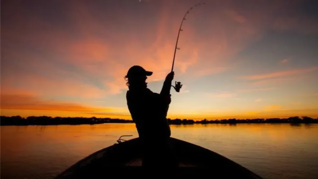 Silueta de una persona con caña de pescar sobre una embarcación al atardecer.